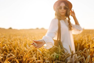 Gün batımında altın buğday kulağına dokunan şapkalı genç bir kadının yakın çekimi. Güzel bir kadın yürümekten zevk alıyor, açık havada özgürlüğü hissediyor. Güzellik ve yürüyüş konsepti.
