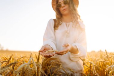 Gün batımında altın buğday kulağına dokunan şapkalı genç bir kadının yakın çekimi. Güzel bir kadın yürümekten zevk alıyor, açık havada özgürlüğü hissediyor. Güzellik ve yürüyüş konsepti.