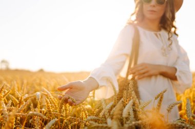 Gün batımında altın buğday kulağına dokunan şapkalı genç bir kadının yakın çekimi. Güzel bir kadın yürümekten zevk alıyor, açık havada özgürlüğü hissediyor. Güzellik ve yürüyüş konsepti.