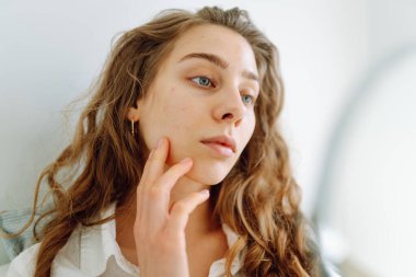 Şirin bir kadın parlak bir odadaki küçük bir aynada kızarıklıklarını inceliyor. Güzel bir kadın güneş ışınlarında doğal güzelliğinin tadını çıkarır. Dermatoloji ve akne araştırması.