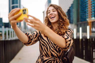 Selfie zamanı! Elinde telefon olan güzel bir kadın arka planda şehirle birlikte gezinti güvertesinde yürüyor. Genç turist blogları ve açık havada selfie çekiyor. Turizm ve blog kavramı.