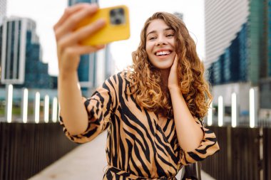 Selfie zamanı! Elinde telefon olan güzel bir kadın arka planda şehirle birlikte gezinti güvertesinde yürüyor. Genç turist blogları ve açık havada selfie çekiyor. Turizm ve blog kavramı.