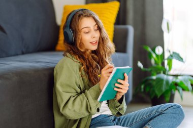 Kablosuz kulaklıklı, elinde kitap tutan genç bir kadın rahat bir odada yerde oturuyor. Bir öğrenci kulaklıkla müzik dinlemekten ve evde çalışmaktan hoşlanır. Eğitim ve konfor kavramı.