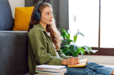 Kablosuz kulaklıklı, elinde kitap tutan genç bir kadın rahat bir odada yerde oturuyor. Bir öğrenci kulaklıkla müzik dinlemekten ve evde çalışmaktan hoşlanır. Eğitim ve konfor kavramı.