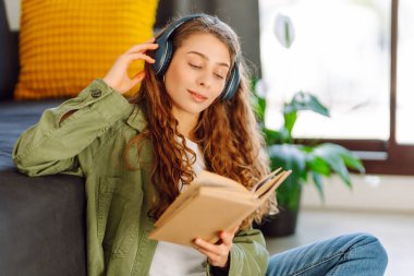 Kablosuz kulaklıklı, elinde kitap tutan genç bir kadın rahat bir odada yerde oturuyor. Bir öğrenci kulaklıkla müzik dinlemekten ve evde çalışmaktan hoşlanır. Eğitim ve konfor kavramı.