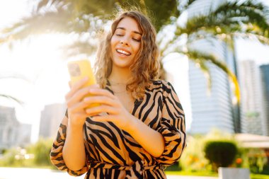 Şehir parkında telefonuyla güneşli bir günün tadını çıkaran mutlu kadın. Telefonda yürüyen, sohbet eden ya da blog yazan genç bir turist. Tatil, hafta sonu ve blog konsepti.