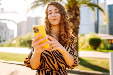 Şehir parkında telefonuyla güneşli bir günün tadını çıkaran mutlu kadın. Telefonda yürüyen, sohbet eden ya da blog yazan genç bir turist. Tatil, hafta sonu ve blog konsepti.