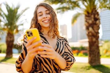 Şehir parkında telefonuyla güneşli bir günün tadını çıkaran mutlu kadın. Telefonda yürüyen, sohbet eden ya da blog yazan genç bir turist. Tatil, hafta sonu ve blog konsepti.