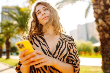Şehir parkında telefonuyla güneşli bir günün tadını çıkaran mutlu kadın. Telefonda yürüyen, sohbet eden ya da blog yazan genç bir turist. Tatil, hafta sonu ve blog konsepti.