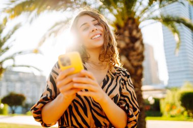 Şehir parkında telefonuyla güneşli bir günün tadını çıkaran mutlu kadın. Telefonda yürüyen, sohbet eden ya da blog yazan genç bir turist. Tatil, hafta sonu ve blog konsepti.