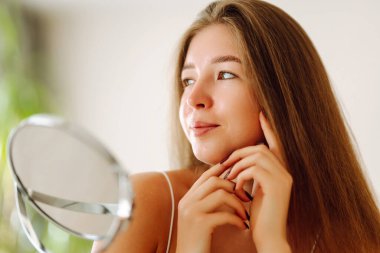 Aynaya bakan sevimli bir kadının portresi. Sıcak bir odada güneşin tadını çıkaran genç bir kadın. Dermatoloji ve güzellik konsepti.