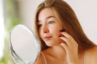 Aynaya bakan sevimli bir kadının portresi. Sıcak bir odada güneşin tadını çıkaran genç bir kadın. Dermatoloji ve güzellik konsepti.