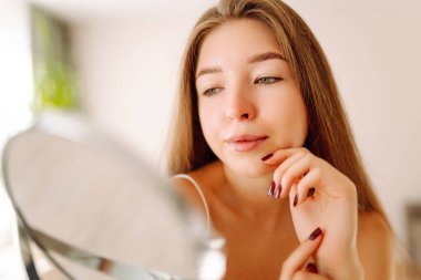 Aynaya bakan sevimli bir kadının portresi. Sıcak bir odada güneşin tadını çıkaran genç bir kadın. Dermatoloji ve güzellik konsepti.