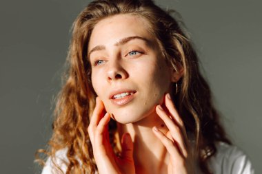 Kıvırcık saçlı genç bir kadın yüzüne hafifçe dokunuyor. Güzel bir kadın sıcak bir odada güneş ışınlarından hoşlanır. Doğallık, güzellik ve rahatlama kavramı..