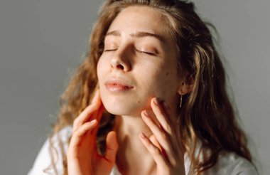 Kıvırcık saçlı genç bir kadın yüzüne hafifçe dokunuyor. Güzel bir kadın sıcak bir odada güneş ışınlarından hoşlanır. Doğallık, güzellik ve rahatlama kavramı..