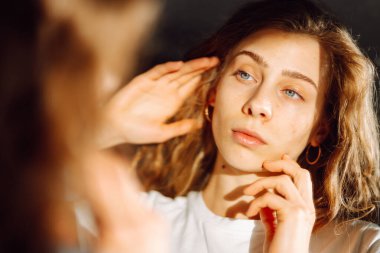 Kıvırcık saçlı genç bir kadın yüzüne hafifçe dokunuyor. Güzel bir kadın sıcak bir odada güneş ışınlarından hoşlanır. Doğallık, güzellik ve rahatlama kavramı..