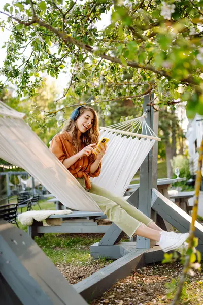 Kablosuz kulaklıklı ve telefonlu şık bir kadın güneşli parkta hamakta uzanıyor. Güzel bir kadın müzik dinlemeyi ya da podcast yapmayı seviyor. Rahatlama ve teknoloji kavramı.