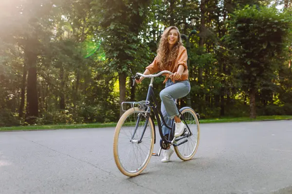 Yeşil bir parkta bisiklet süren genç bir kadın. Neşeli kadın hafta sonunun tadını çıkarıyor, gün batımında bisiklet sürüyor. Aktif yaşam tarzı. Joy, hafta sonu konsepti.