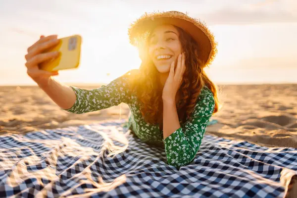 Hasır şapkalı ve parlak elbiseli neşeli bir kadın deniz kenarındaki bir paspasın üzerinde yatarken telefonunda selfie çekiyor. Güzel bir turist deniz manzarası ve bloglardan hoşlanır. Bloglama, hafta sonu konsepti.