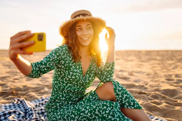 Hasır şapkalı ve parlak elbiseli neşeli bir kadın deniz kenarındaki bir paspasın üzerinde yatarken telefonunda selfie çekiyor. Güzel bir turist deniz manzarası ve bloglardan hoşlanır. Bloglama, hafta sonu konsepti.