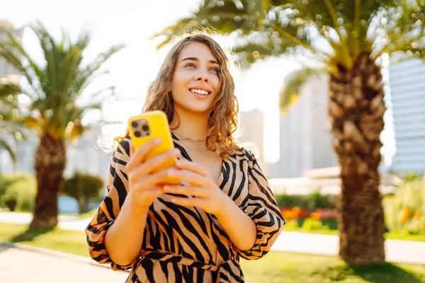 Şehir parkında telefonuyla güneşli bir günün tadını çıkaran mutlu kadın. Telefonda yürüyen, sohbet eden ya da blog yazan genç bir turist. Tatil, hafta sonu ve blog konsepti.