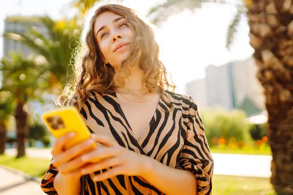 Şehir parkında telefonuyla güneşli bir günün tadını çıkaran mutlu kadın. Telefonda yürüyen, sohbet eden ya da blog yazan genç bir turist. Tatil, hafta sonu ve blog konsepti.