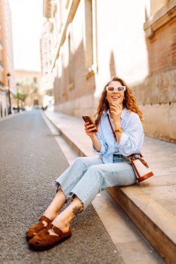 Güneş gözlüğü takan, elinde telefon tutan, eski kasabada yürüyüş yapmaktan zevk alan zarif genç bir kadın. Yürüyüş, turizm, teknoloji konsepti. Açık havada dinlenirken telefonla turist blogları.