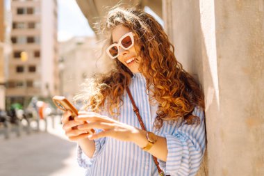 Güneş gözlüğü takan, elinde telefon tutan, eski kasabada yürüyüş yapmaktan zevk alan zarif genç bir kadın. Yürüyüş, turizm, teknoloji konsepti. Açık havada dinlenirken telefonla turist blogları.