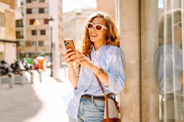 Güneş gözlüğü takan, elinde telefon tutan, eski kasabada yürüyüş yapmaktan zevk alan zarif genç bir kadın. Yürüyüş, turizm, teknoloji konsepti. Açık havada dinlenirken telefonla turist blogları.