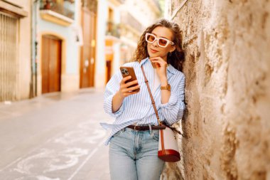 Güneş gözlüğü takan, elinde telefon tutan, eski kasabada yürüyüş yapmaktan zevk alan zarif genç bir kadın. Yürüyüş, turizm, teknoloji konsepti. Açık havada dinlenirken telefonla turist blogları.