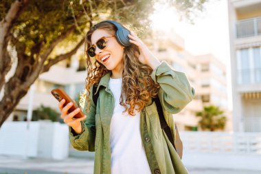 Güneş gözlüklü, telefon ve kablosuz kulaklıklı şık bir kadın güneşli parkta havanın tadını çıkarıyor. Açık deniz kenarında yürüyen güzel bir turist. Teknoloji ve keyif kavramı.