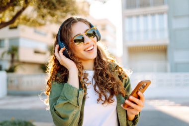 Güneş gözlüklü, telefon ve kablosuz kulaklıklı şık bir kadın güneşli parkta havanın tadını çıkarıyor. Açık deniz kenarında yürüyen güzel bir turist. Teknoloji ve keyif kavramı.