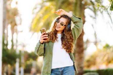 Güneş gözlüklü, telefon ve kablosuz kulaklıklı şık bir kadın güneşli parkta havanın tadını çıkarıyor. Açık deniz kenarında yürüyen güzel bir turist. Teknoloji ve keyif kavramı.