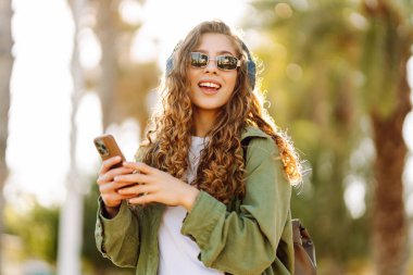 Güneş gözlüklü, telefon ve kablosuz kulaklıklı şık bir kadın güneşli parkta havanın tadını çıkarıyor. Açık deniz kenarında yürüyen güzel bir turist. Teknoloji ve keyif kavramı.