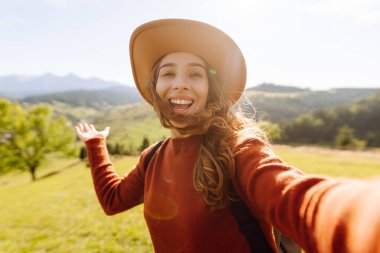 Telefonuyla selfie çeken şapkalı bir gezginin portresi. Neşeli bir kadın dağ manzarasını ve blogları seviyor. Seyahat, günlük tutma kavramı.