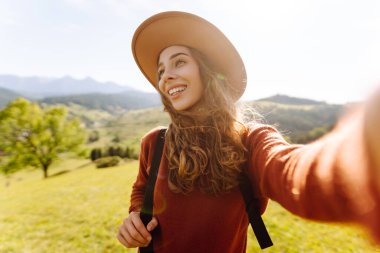 Telefonuyla selfie çeken şapkalı bir gezginin portresi. Neşeli bir kadın dağ manzarasını ve blogları seviyor. Seyahat, günlük tutma kavramı.