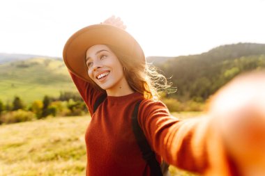 Telefonuyla selfie çeken şapkalı bir gezginin portresi. Neşeli bir kadın dağ manzarasını ve blogları seviyor. Seyahat, günlük tutma kavramı.