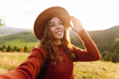 Telefonuyla selfie çeken şapkalı bir gezginin portresi. Neşeli bir kadın dağ manzarasını ve blogları seviyor. Seyahat, günlük tutma kavramı.