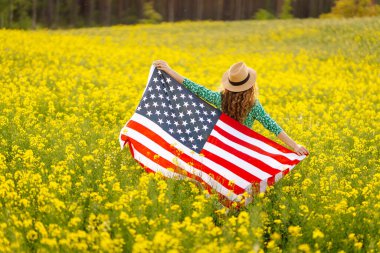 Parlak sarı bir tarlada neşeli bir kadın güneşli bir günde Birleşik Devletler bayrağını tutuyor. Güzel bir kadın özgürlüğü hisseder, kolza tohumu tarlasından zevk alır. Tatil kavramı, eğlence..