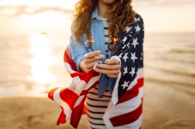 Amerikan bayrağına sarılı genç bir kadın gün batımı kumsalında maytap tutuyor. Gün batımının tadını çıkaran güzel kadın. Bağımsızlık kavramı, kutlama.
