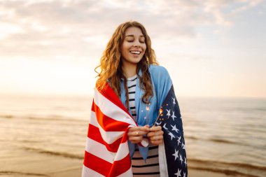 Amerikan bayrağına sarılı genç bir kadın gün batımı kumsalında maytap tutuyor. Gün batımının tadını çıkaran güzel kadın. Bağımsızlık kavramı, kutlama.