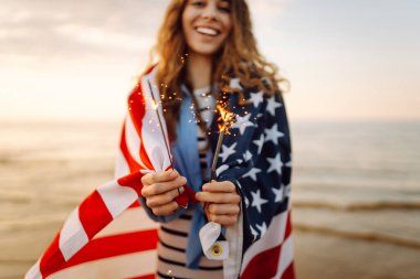 Amerikan bayrağına sarılı genç bir kadın gün batımı kumsalında maytap tutuyor. Gün batımının tadını çıkaran güzel kadın. Bağımsızlık kavramı, kutlama.