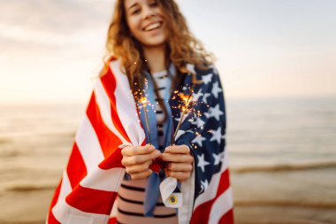 Amerikan bayrağına sarılı genç bir kadın gün batımı kumsalında maytap tutuyor. Gün batımının tadını çıkaran güzel kadın. Bağımsızlık kavramı, kutlama.