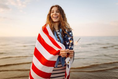 Amerikan bayrağına sarılı genç bir kadın gün batımı kumsalında maytap tutuyor. Gün batımının tadını çıkaran güzel kadın. Bağımsızlık kavramı, kutlama.