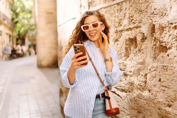Güneş gözlüğü takan, elinde telefon tutan, eski kasabada yürüyüş yapmaktan zevk alan zarif genç bir kadın. Yürüyüş, turizm, teknoloji konsepti. Açık havada dinlenirken telefonla turist blogları.