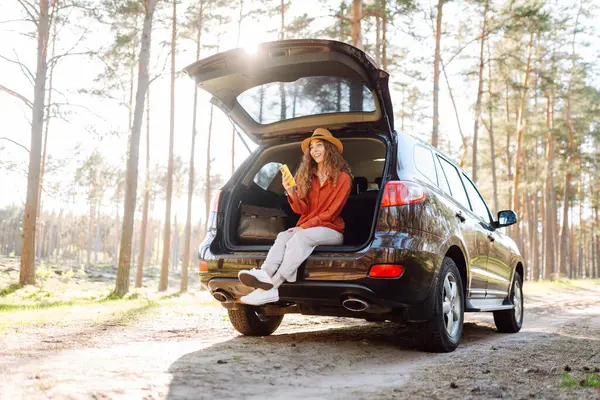 Gözlüklü ve telefonlu güzel bir kadın bir arabanın bagajında oturuyor ve günbatımı ormanının tadını çıkarıyor. Yolculukta olan genç bir kadın dışarıda blog yazıyor. Macera, blog konsepti.