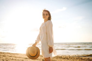 Güneşli sahilde yürüyen, şapkalı ve güneş gözlüklü gülümseyen bir kadın. Güzel bir kadın sahilde yürümekten ve özgürlüğü hissetmekten zevk alıyor. Rahatlama ve keyif kavramı.