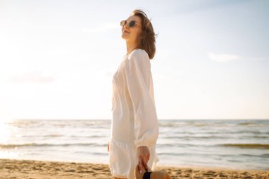 Güneşli sahilde yürüyen, şapkalı ve güneş gözlüklü gülümseyen bir kadın. Güzel bir kadın sahilde yürümekten ve özgürlüğü hissetmekten zevk alıyor. Rahatlama ve keyif kavramı.