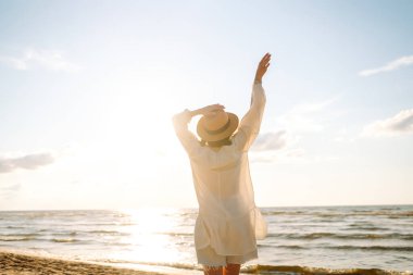 Güneşli sahilde yürüyen, şapkalı ve güneş gözlüklü gülümseyen bir kadın. Güzel bir kadın sahilde yürümekten ve özgürlüğü hissetmekten zevk alıyor. Rahatlama ve keyif kavramı.