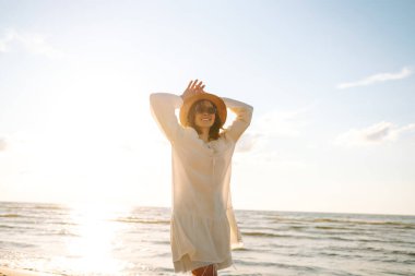 Güneşli sahilde yürüyen, şapkalı ve güneş gözlüklü gülümseyen bir kadın. Güzel bir kadın sahilde yürümekten ve özgürlüğü hissetmekten zevk alıyor. Rahatlama ve keyif kavramı.